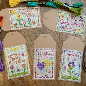 以下が含まれることがあります： 茶色の厚紙に手作りのクロスステッチで作った「お大事に」カードが5枚。カードには、花、太陽、風船など、さまざまなデザインが施されています。カードに書かれている言葉は、「Get Well Soon」、「Better Days Ahead」、「Thinking of You」、「Get Well Soon」です。