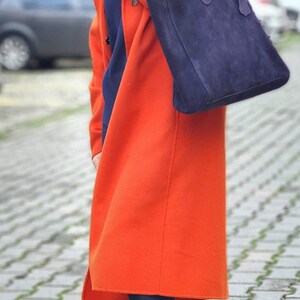 Op de afbeelding: Een lange, feloranje jas en een marineblauwe su&egrave;de tote bag. De jas is knielang en heeft een klassiek ontwerp. De tas wordt over de schouder gedragen. De persoon draagt een donkerblauwe jeans en kleurrijke sneakers.