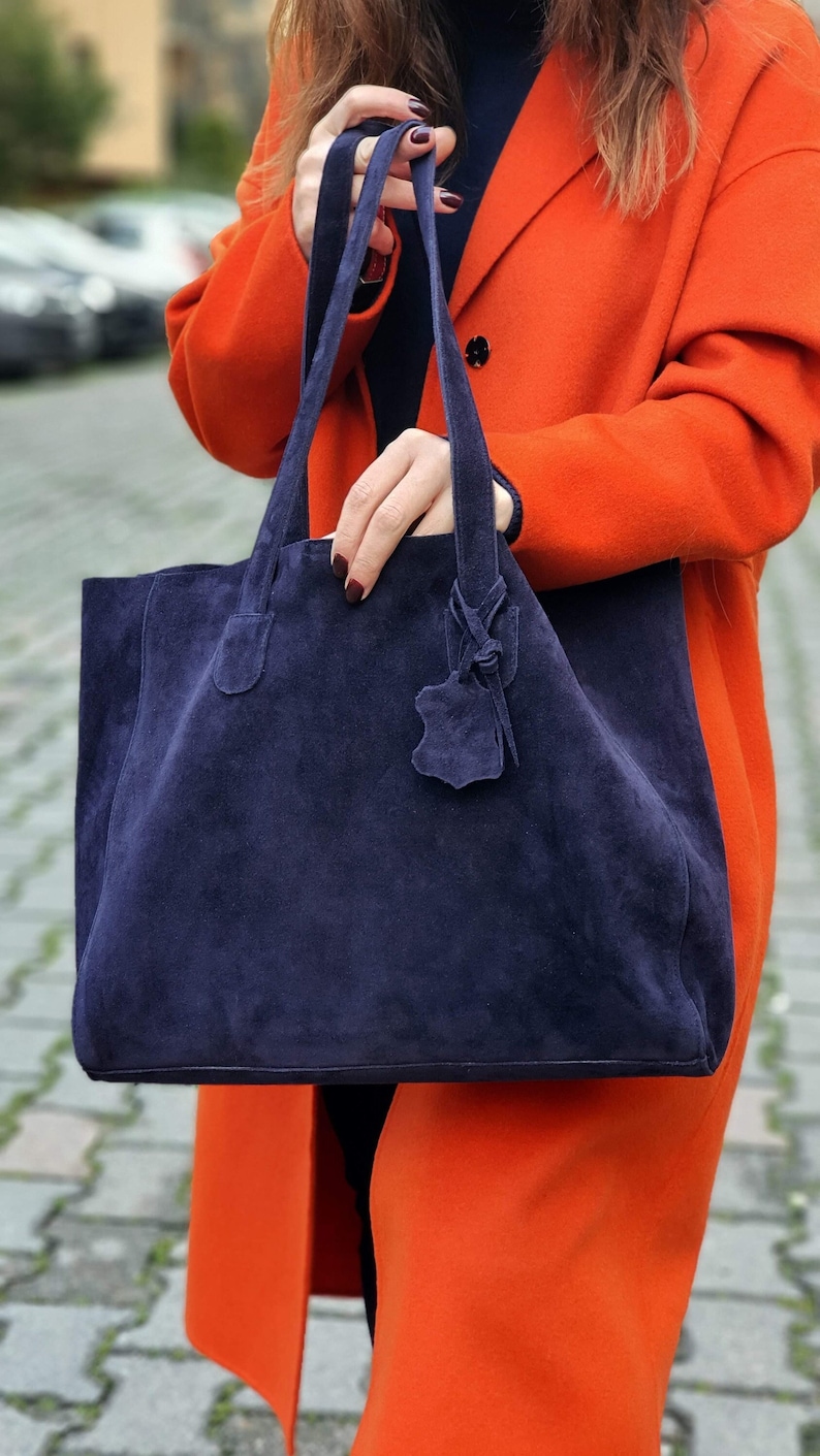 Op de afbeelding: Een marineblauwe su&egrave;de tote bag met lange handvatten. De tas heeft een rechthoekige vorm en een klein leren label aan het handvat. De persoon draagt een oranje jas.