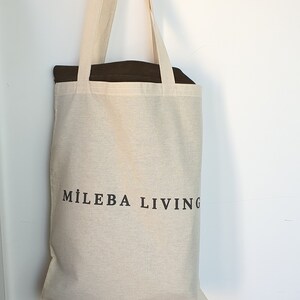 Op de afbeelding: Een beige canvas draagtas met de tekst "MILEBA LIVING" in zwart gedrukt. De tas heeft lange handvatten en hangt tegen een witte muur. De binnenkant van de tas is donkerbruin.