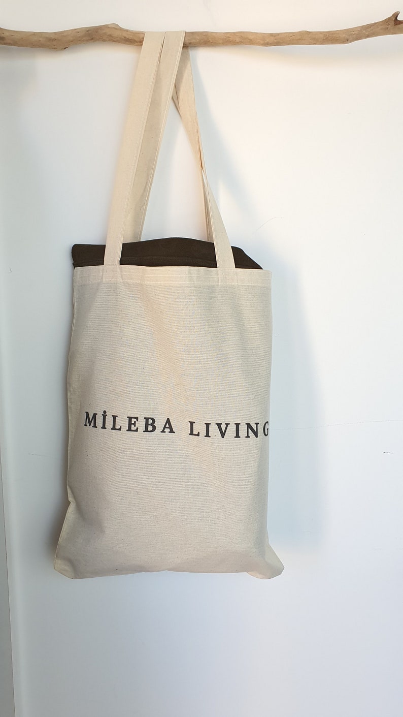 Op de afbeelding: Een beige canvas draagtas met de tekst "MILEBA LIVING" in zwart gedrukt. De tas heeft lange handvatten en hangt tegen een witte muur. De binnenkant van de tas is donkerbruin.
