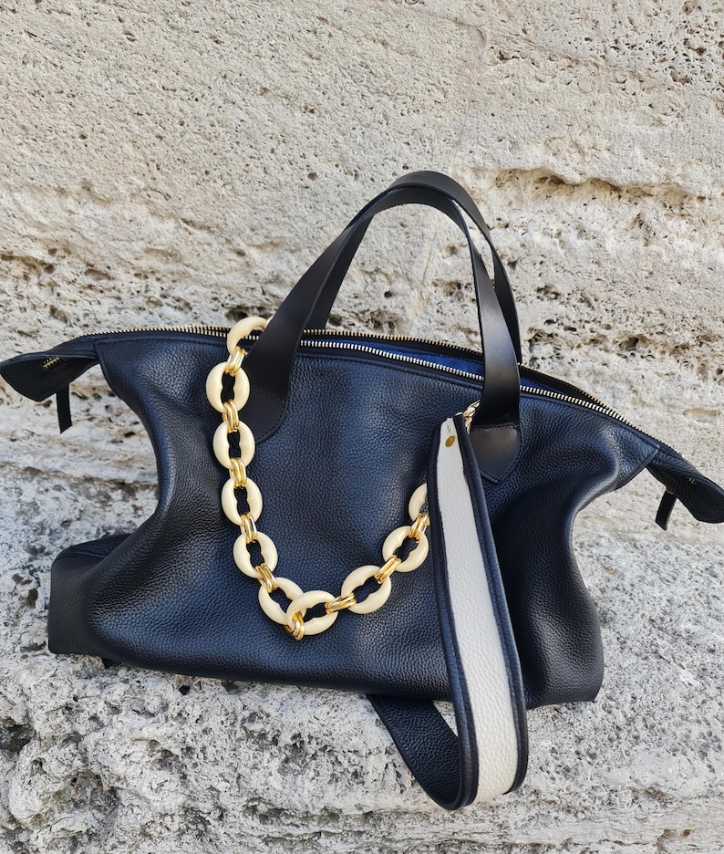 May include: Black leather tote bag with a gold chain strap and a white and black leather strap. The bag has a zipper closure and a top handle.