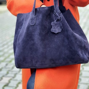 Op de afbeelding: Een grote, marineblauwe su&egrave;de tote bag met lange handvatten en een leren label. De tas wordt vastgehouden tegen een feloranje jas. De jas en tas cre&euml;ren een opvallend kleurcontrast. De tas lijkt ruim.