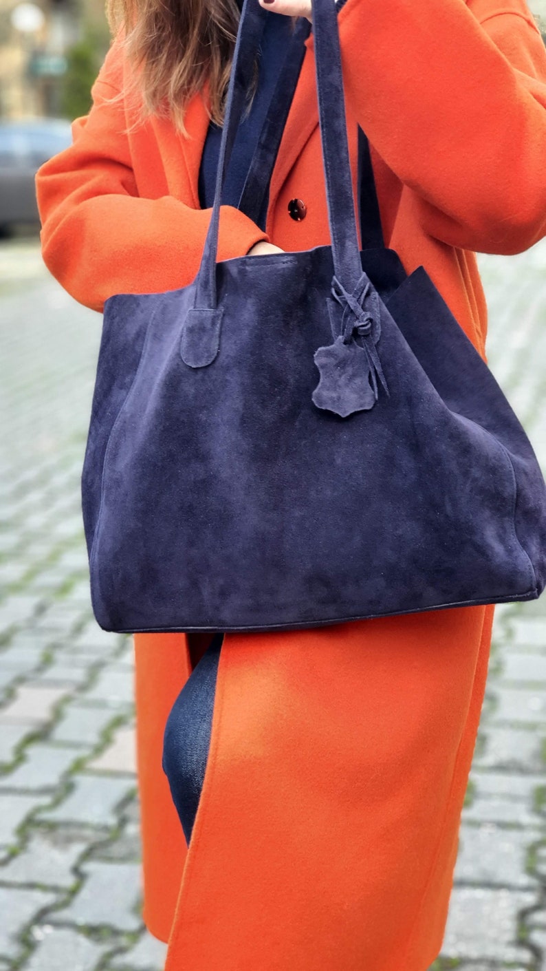Op de afbeelding: Een grote, marineblauwe su&egrave;de tote bag met lange handvatten en een leren label. De tas wordt vastgehouden tegen een feloranje jas. De jas en tas cre&euml;ren een opvallend kleurcontrast. De tas lijkt ruim.