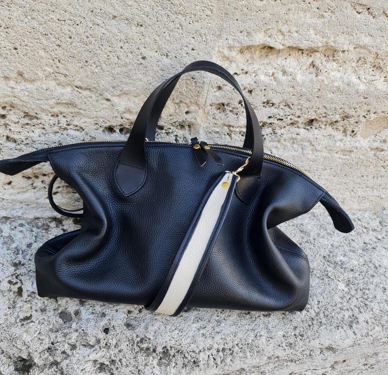 May include: A black leather tote bag with a white and black strap. The bag has a zipper closure and gold hardware.