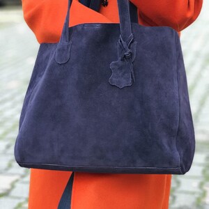 Op de afbeelding: Een grote, marineblauwe su&egrave;de tote bag met bijpassende handvatten en een decoratief leren label. De tas wordt vastgehouden tegen een achtergrond van een feloranje jas. De zachte textuur van de tas contrasteert met de gladde stof van de jas.