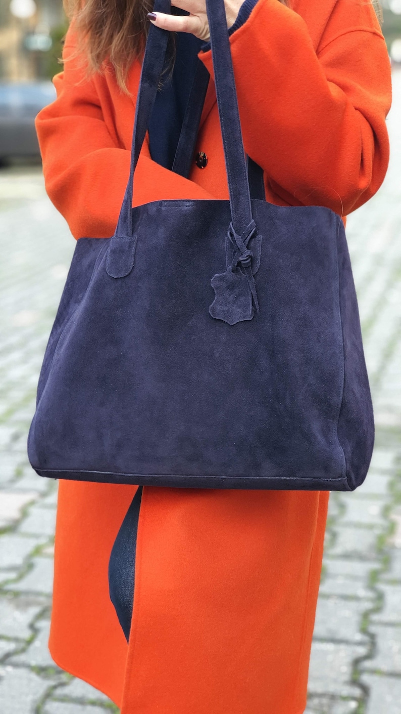 Op de afbeelding: Een grote, marineblauwe su&egrave;de tote bag met bijpassende handvatten en een decoratief leren label. De tas wordt vastgehouden tegen een achtergrond van een feloranje jas. De zachte textuur van de tas contrasteert met de gladde stof van de jas.