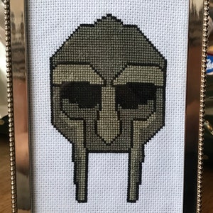 May include: A framed cross-stitch of a gray and black mask with a geometric design. The mask is in a silver frame with a beaded border.