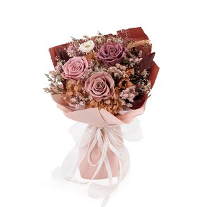 Op de afbeelding: Een boeket van geconserveerde bloemen met drie grote rozen in tinten oudroze en mauve, geaccentueerd met gedroogd gipskruid, beige hortensia's en donkerbordeauxrode bladeren. Het arrangement is verpakt in roze papier en gestrikt met een wit lint.