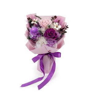 Op de afbeelding: Een bloemenboeket met een grote lichtroze roos, een dieppaarse roos en een getextureerde paarse bloem. Het arrangement bevat witte vulbloemen, donkergroen blad en roze gedroogde grassen, verpakt in lichtroze papier met een paars lint.