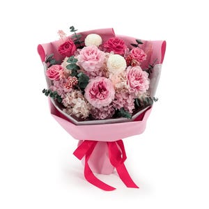 Op de afbeelding: Een boeket van geconserveerde bloemen met diverse tinten roze rozen, witte bloesems en groene eucalyptusbladeren. Het arrangement is verpakt in lichtroze papier en vastgebonden met een felroze lint, wat zorgt voor een gestructureerd bloemstuk.