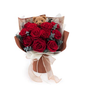 Op de afbeelding: Een boeket van dieprode rozen, geschikt met kleine groene eucalyptus takjes en een kleine bruine pluche teddybeer in het midden. De bloemen zijn verpakt in bruin en doorschijnend wit papier, vastgebonden met een beige lint.