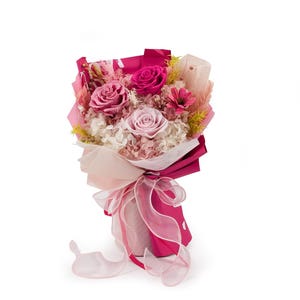 Op de afbeelding: Een bloemenboeket met geconserveerde rozen in tinten diep magenta, mauve en lichtroze, geaccentueerd met witte hortensia's, gedroogde grassen en kleine gele takjes. Het arrangement is verpakt in magenta en doorschijnend papier met een lint.