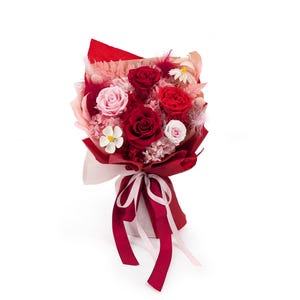 Op de afbeelding: Een bloemenboeket met donkerrode en zachtroze rozen, geaccentueerd met kleine witte bloemetjes, gedroogd blad en fijne veertjes. Het arrangement is verpakt in rood en beige papier en samengebonden met een lang, tweekleurig roze en rood lint.