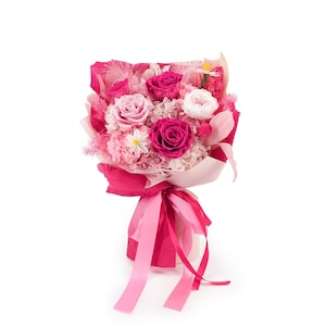 Op de afbeelding: Een bloemenboeket met diepmagenta en lichtroze rozen, witte vulbloemen en kleine madeliefachtige bloesems. Het arrangement is verpakt in levendig magenta en lichtroze papier, vastgezet met lange, zwierige roze satijnen linten.