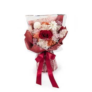 Op de afbeelding: Een boeket van geconserveerde bloemen met een centrale dieprode roos omringd door witte, oranje en lichtroze bloesems, gedroogde grassen en kleine paarse accenten. Het arrangement is verpakt in doorschijnend en donkerrood papier met een satijnen lint.