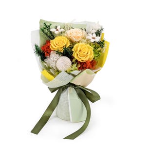 Op de afbeelding: Een boeket van geconserveerde bloemen met twee opvallende gele rozen, een licht perzikkleurige roos, witte vulbloemen en groen blad. Het arrangement is verpakt in lichtgroen en geel papier, vastgebonden met een donker olijfgroen lint.