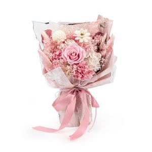 Op de afbeelding: Een boeket van geconserveerde bloemen met een grote roze roos in het midden, omringd door witte en roze droogbloemen, pluizig pampasgras en kleine witte vulbloemen. Het arrangement is verpakt in roze papier met een roze satijnen lint.