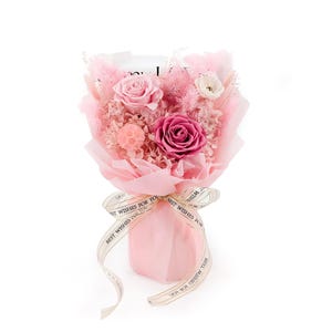 Op de afbeelding: Een boeket van geconserveerde bloemen met een lichtroze roos, een diepmagenta roos en kleine witte bloesems, geaccentueerd met roze pluizig gras. Het arrangement is verpakt in doorschijnend roze papier en gestrikt met een lint waarop de tekst BEST WISHES FOR YOU staat.