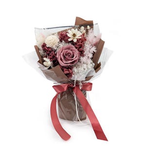 Op de afbeelding: Een boeket van geconserveerde bloemen met een grote oudroze centrale roos, witte en bordeauxrode accenten en vederachtige roze pluimen. Het arrangement is verpakt in bruin papier en doorzichtig plastic, gestrikt met een donkerrood satijnen lint.