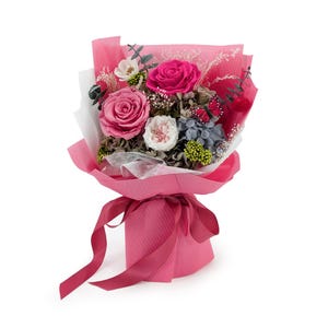 Op de afbeelding: Een boeket van geconserveerde bloemen met twee grote roze rozen, een kleinere witte roos, blauwe hortensia's, groene bessen en eucalyptus takjes. Een decoratieve vlinder is toegevoegd. Het boeket is verpakt in roze papier met een strik.