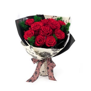 Op de afbeelding: Een boeket van levendige rode rozen met groen blad, verpakt in crèmekleurig papier met zwarte bloemmotieven en tekst, vastgebonden met een decoratief bruin lint bedrukt met een donker bloemmotief op een witte achtergrond.