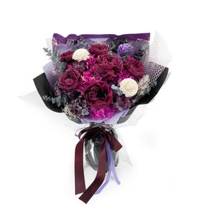 Op de afbeelding: Een boeket van diep bordeauxrode en paarse rozen gemengd met witte bloemen, paarse hortensia's en groen eucalyptusblad. Het arrangement is verpakt in zwart en paars papier met een decoratieve netlaag en vastgebonden met een donker lint.