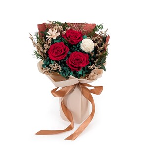 Op de afbeelding: Een bloemenboeket met drie levendige rode rozen, twee kleine witte bloemen en groen blad. Het arrangement is verpakt in beige papier met een textuur van roodbruine achtergrond en vastgebonden met een lang, glad, lichtbruin satijnen lint.