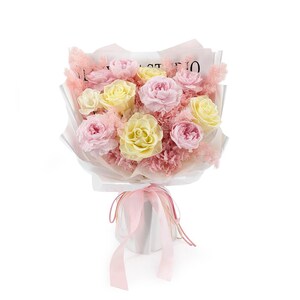Op de afbeelding: Een boeket van geconserveerde lichtroze en crèmekleurige rozen, geschikt met zachtroze, vederachtige vulbloemen. Het arrangement is verpakt in wit papier met de tekst STUDIO zichtbaar aan de bovenkant en gestrikt met een lichtroze lint.