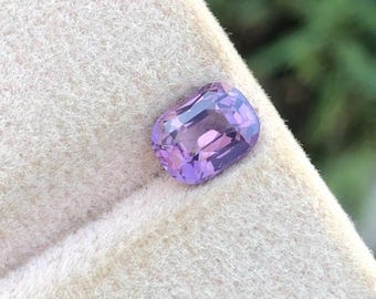 Può includere: Una gemma sfaccettata, taglio rettangolare, con una sfumatura di tonalità viola. La gemma è posta su una superficie strutturata color crema, con uno sfondo verde sfocato. Le sfaccettature della gemma riflettono la luce, mostrando la sua chiarezza.
