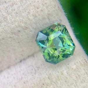 Puede incluir: Una gema facetada, de talla esmeralda, con un tono verde vibrante. La gema muestra una gama de tonos verdes, de claros a oscuros, con una superficie pulida. La gema se sostiene sobre un fondo blanco texturizado.
