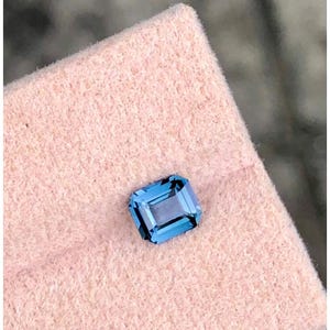 May include: A rectangular, light blue gemstone with a step cut. The gem is set on a pink, textured surface. The gemstone has a bright, reflective quality.