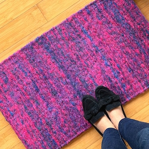 May include: A pink and purple woven rug with a fuzzy texture. Two black suede loafers are on top of the rug.