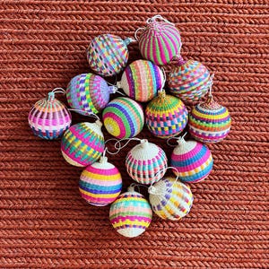 May include: A collection of colorful, woven sphere ornaments with white string loops. The ornaments feature vibrant patterns of pink, green, blue, yellow, and purple. The background is a textured, reddish-brown surface.