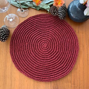 May include: A round, burgundy-colored placemat with a spiral design. The placemat is made of a woven material and is placed on a wooden surface. The image also includes decorative elements such as pinecones, flowers, and glassware.
