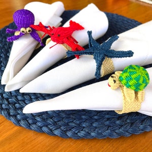 May include: Four white napkins with different sea-themed napkin rings. The rings are a purple octopus, a red lobster, a blue starfish, and a green and yellow turtle.