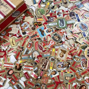 Könnte beinhalten: Eine Sammlung alter Zigarrenetiketten mit verschiedenen Porträts, Illustrationen und Texten. Die Etiketten sind in verschiedenen Formen und Größen gehalten, mit einer Mischung aus Farben wie Rot, Gold und Braun. Eine Holzkiste mit Text ist oben links sichtbar.
