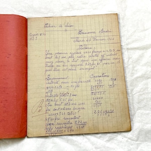 May include: An open vintage ledger with handwritten calculations and French text. The red-covered notebook features lined pages filled with equations and financial records. The handwriting is in dark ink, with the date "Mardi 23 Fevrier 1926" visible.