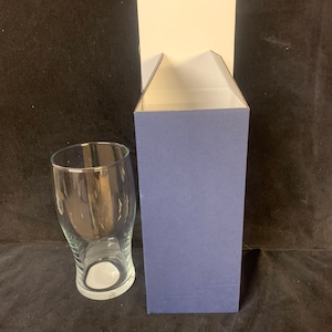 May include: A clear glass beer pint glass with a curved design, next to a blue and white cardboard box. The glass has a wide top and a narrow base. The box is open, revealing a white interior.