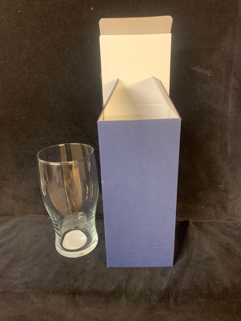 May include: A clear glass beer pint glass with a curved design, next to a blue and white cardboard box. The glass has a wide top and a narrow base. The box is open, revealing a white interior.