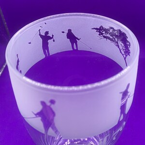 May include: A frosted glass with a golf-themed design. The glass features silhouettes of golfers in action, along with a tree. The base of the glass has a decorative etched design. The background is a solid purple.