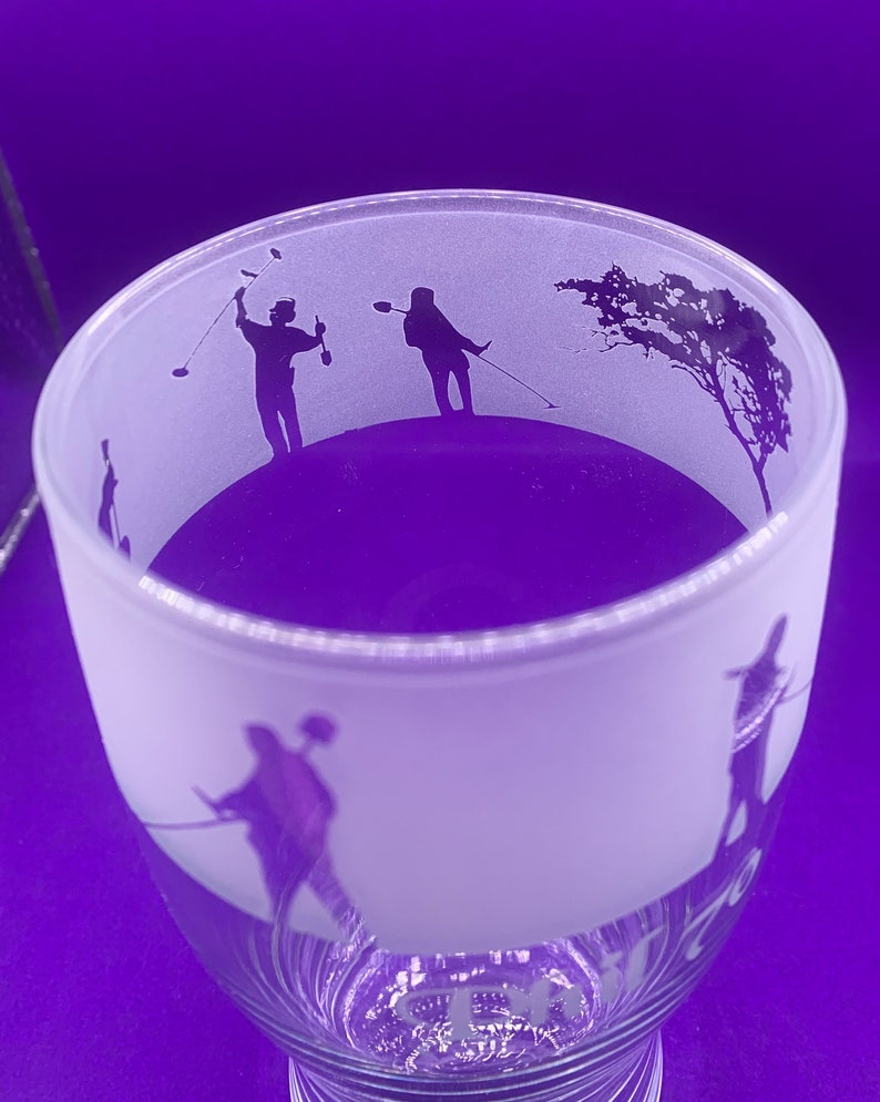 May include: A frosted glass with a golf-themed design. The glass features silhouettes of golfers in action, along with a tree. The base of the glass has a decorative etched design. The background is a solid purple.