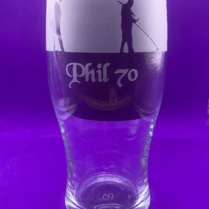 May include: Clear glass pint glass with a frosted top section. The top features silhouettes of people metal detecting. The text "Phil 70" is etched below the frosted section. The glass has a wide top and tapers down.