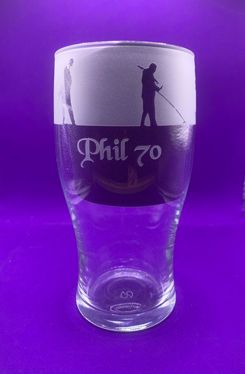 May include: Clear glass pint glass with a frosted top section. The top features silhouettes of people metal detecting. The text "Phil 70" is etched below the frosted section. The glass has a wide top and tapers down.