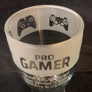 May include: A frosted glass with the text "PRO GAMER" in black and grey lettering. The glass also has two black and white images of video game controllers. The text "Level 46 Achieved Love From Dylan Bethany & Spencer" is written in a smaller font at the bottom of the glass.