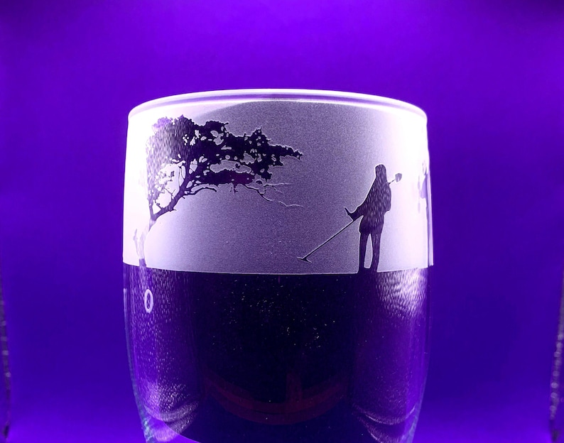 May include: A clear glass with a frosted design featuring a silhouette of a person using a metal detector near a tree. The glass has a purple background and a dark purple base.