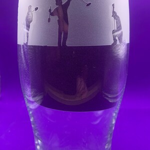 May include: A clear glass beer pint with a frosted band featuring etched silhouettes of three people metal detecting. The figures are in various poses, with one raising a detector. The bottom half of the glass is clear.