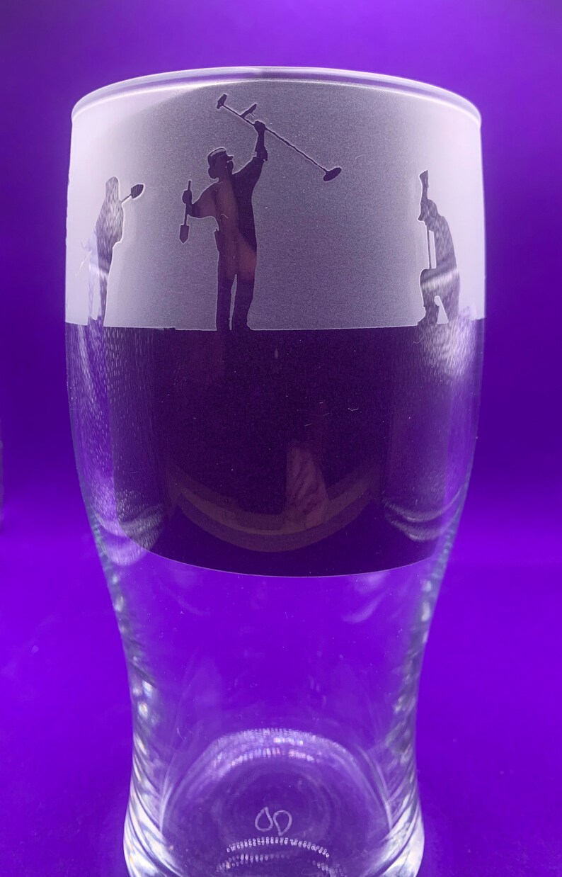 May include: A clear glass beer pint with a frosted band featuring etched silhouettes of three people metal detecting. The figures are in various poses, with one raising a detector. The bottom half of the glass is clear.