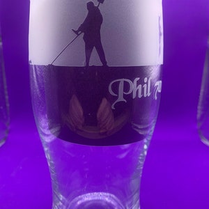 May include: A clear glass beer pint with a frosted top section featuring a silhouette of a person holding a shovel. The glass has the name "Phil" in a decorative font. The glass is on a purple background.