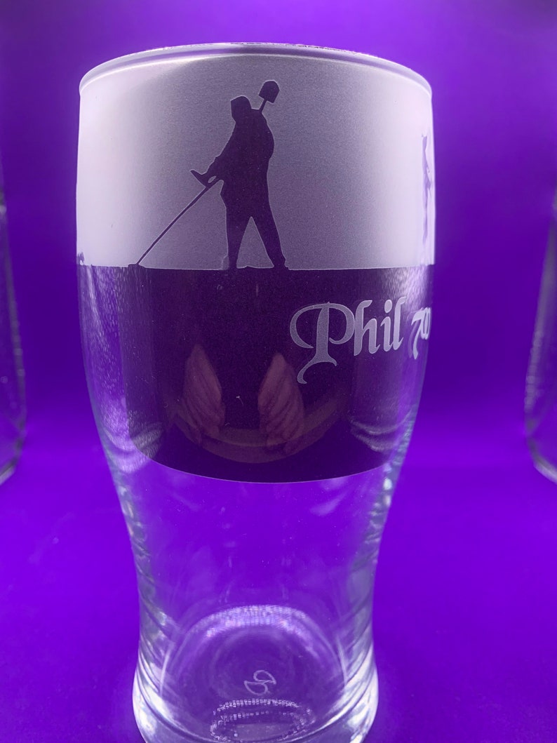 May include: A clear glass beer pint with a frosted top section featuring a silhouette of a person holding a shovel. The glass has the name "Phil" in a decorative font. The glass is on a purple background.